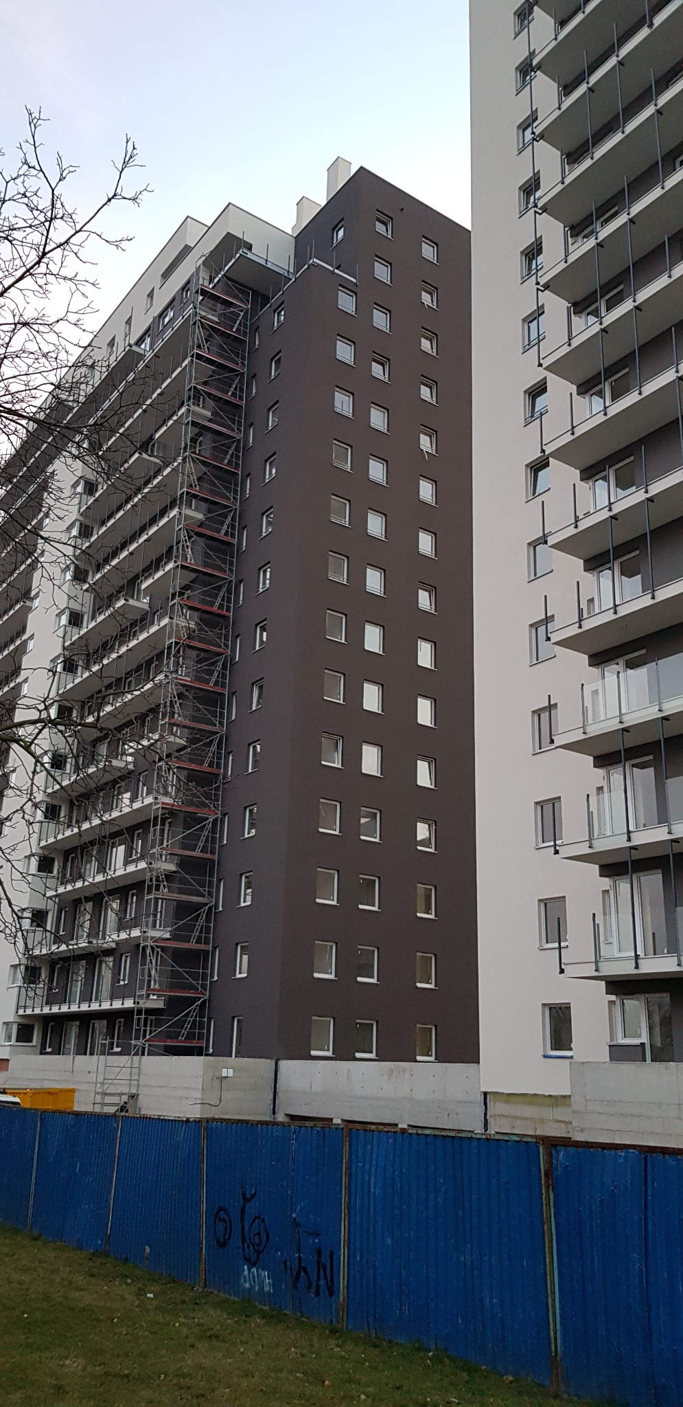 Wielopiętrowy budynek w trakcie prac elewacyjnych, widoczne rusztowania na bocznej ścianie, obok budynek z balkonami, całość odgrodzona niebieskim płotem z graffiti.