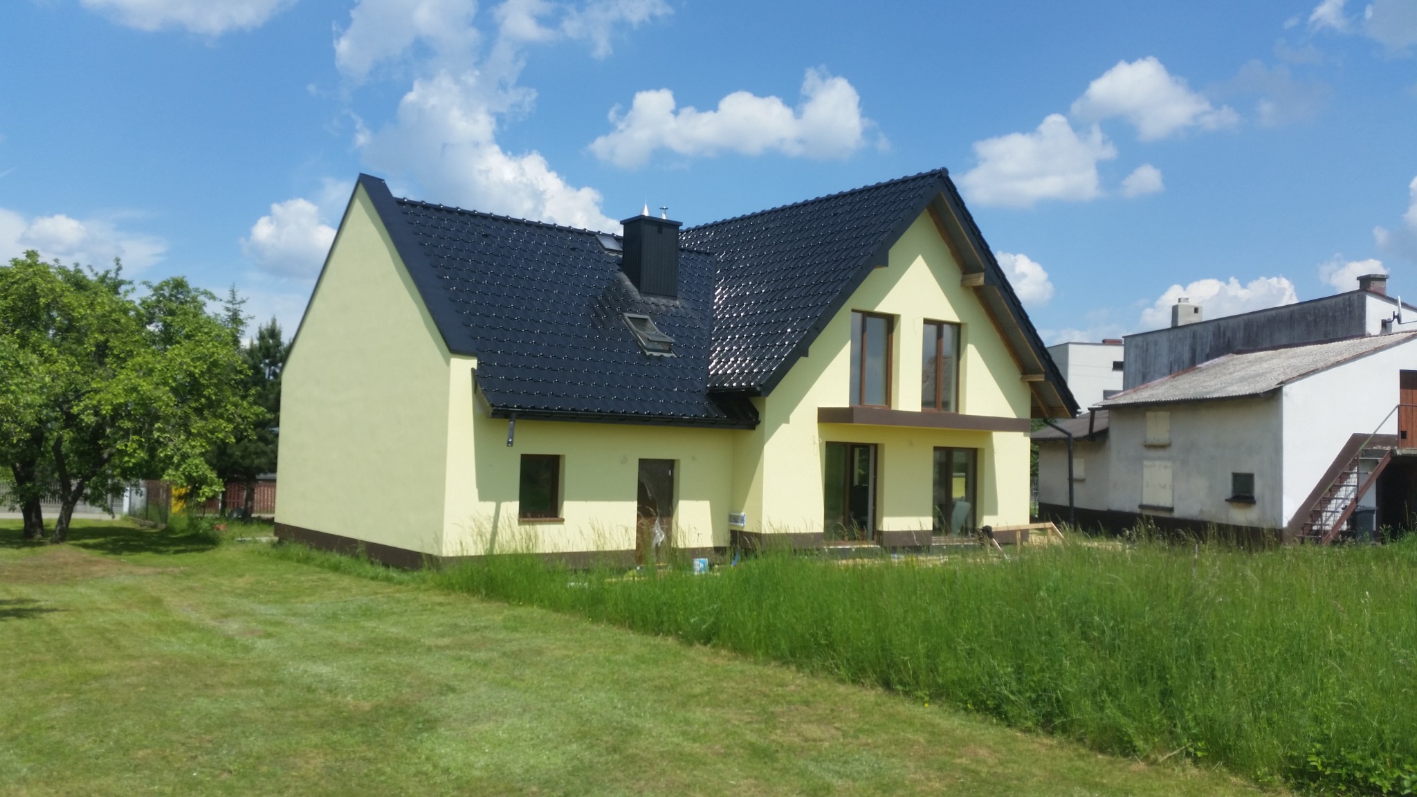 Widok z zewnątrz na dom jednorodzinny z jasną elewacją, ciemną dachówką i zadbanym trawnikiem w słoneczny dzień.