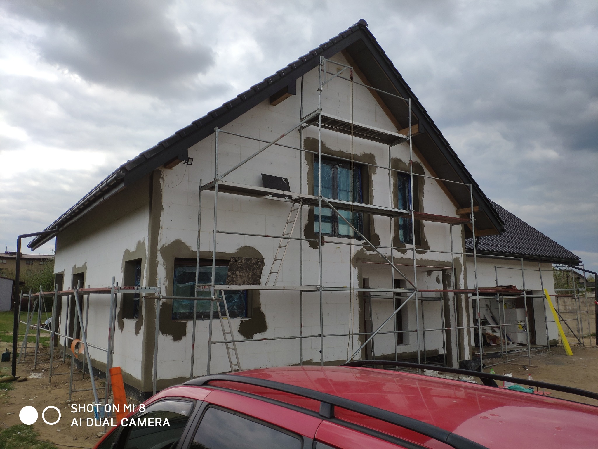 Dom w trakcie ocieplania elewacji, widoczne rusztowania, częściowo otynkowane ściany, zabezpieczone okna folią, fragment czerwonego samochodu na pierwszym planie.
