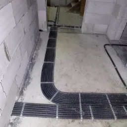 Wiązka przewodów elektrycznych w karbowanej osłonie, przymocowana metalowymi uchwytami do surowej betonowej podłogi w nowym budynku z białego betonu komórkowego.