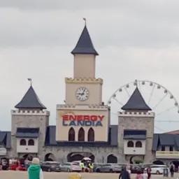 Fasada budynku z wieżą zegarową i napisem Energylandia, w tle diabelski młyn i spacerujący ludzie.