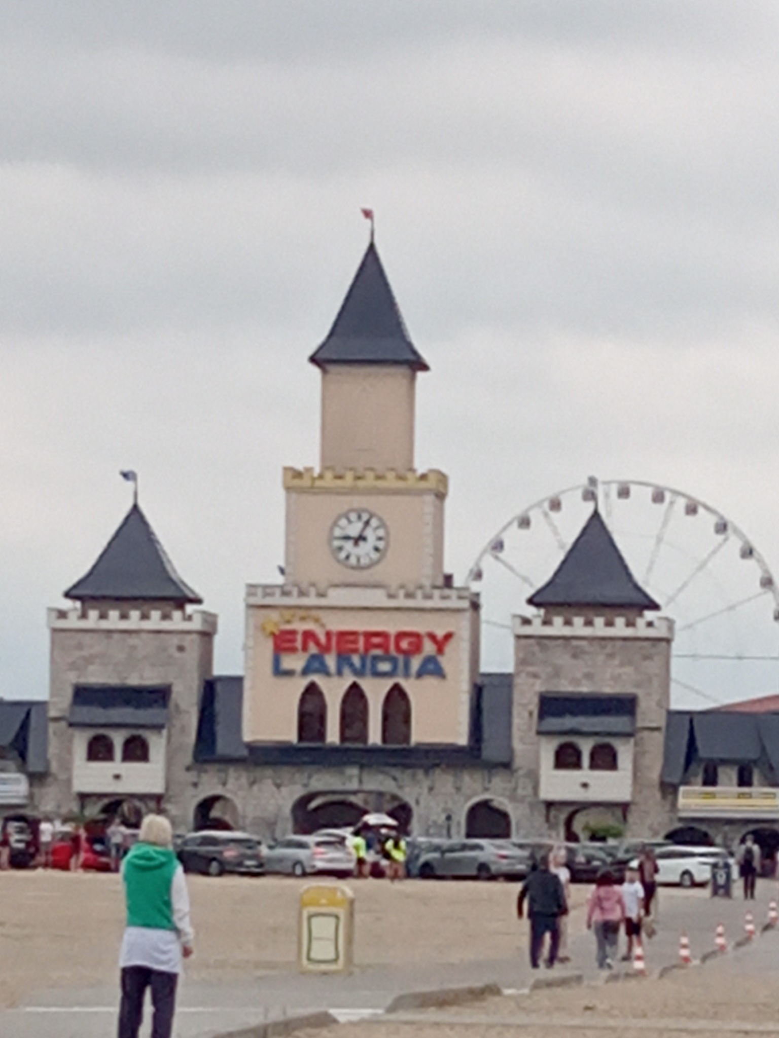 Fasada budynku z wieżą zegarową i napisem Energylandia, w tle diabelski młyn i spacerujący ludzie.