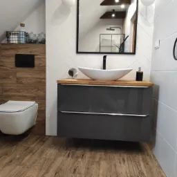 Nowoczesna łazienka na poddaszu z drewnianą podłogą i ścianą, białe płytki ułożone w jodełkę, szara szafka pod umywalką z drewnianym blatem, wisząca toaleta i okrągłe lustro do makijażu.