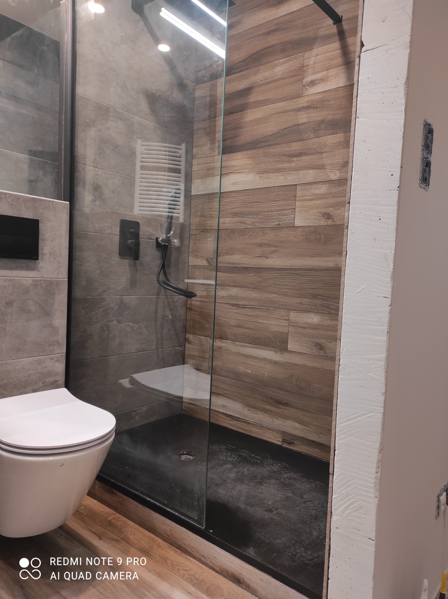 Nowoczesna kabina prysznicowa walk-in z drewnopodobnymi płytkami na ścianie, szklana przegroda, biała toaleta i grzejnik drabinkowy w tle.