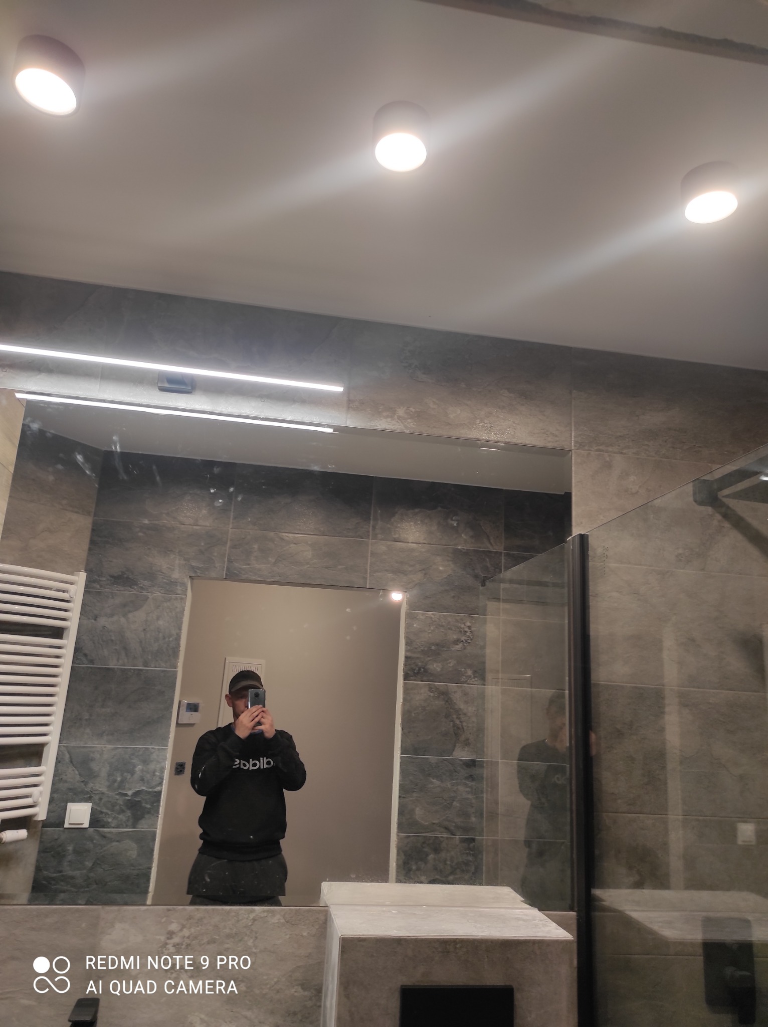 Wykończona łazienka w Starogardzie Gdańskim z szarymi płytkami imitującymi kamień, oświetleniem LED i grzejnikiem drabinkowym, sfotografowana w lustrze przez wykonawcę.