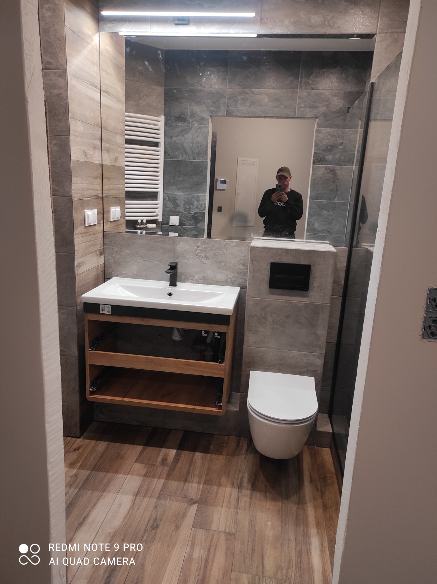Nowoczesna łazienka z drewnopodobnymi płytkami na podłodze i ścianach, umywalką z szafką z otwartymi półkami, toaletą podwieszaną i dużym lustrem, w którym odbija się osoba robiąca zdjęcie.