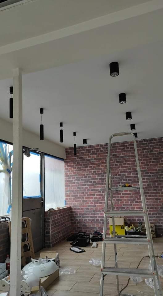 Montaż oświetlenia sufitowego w łazience z ceglaną ścianą, widoczna drabina i materiały budowlane.