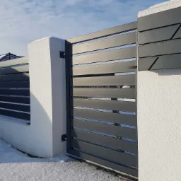 Nowoczesna, pozioma brama wjazdowa z metalowymi panelami w kolorze grafitowym, zamontowana na białych, otynkowanych słupkach. Po prawej stronie, na słupku, skrzynka na listy z geometrycznym wzorem.
