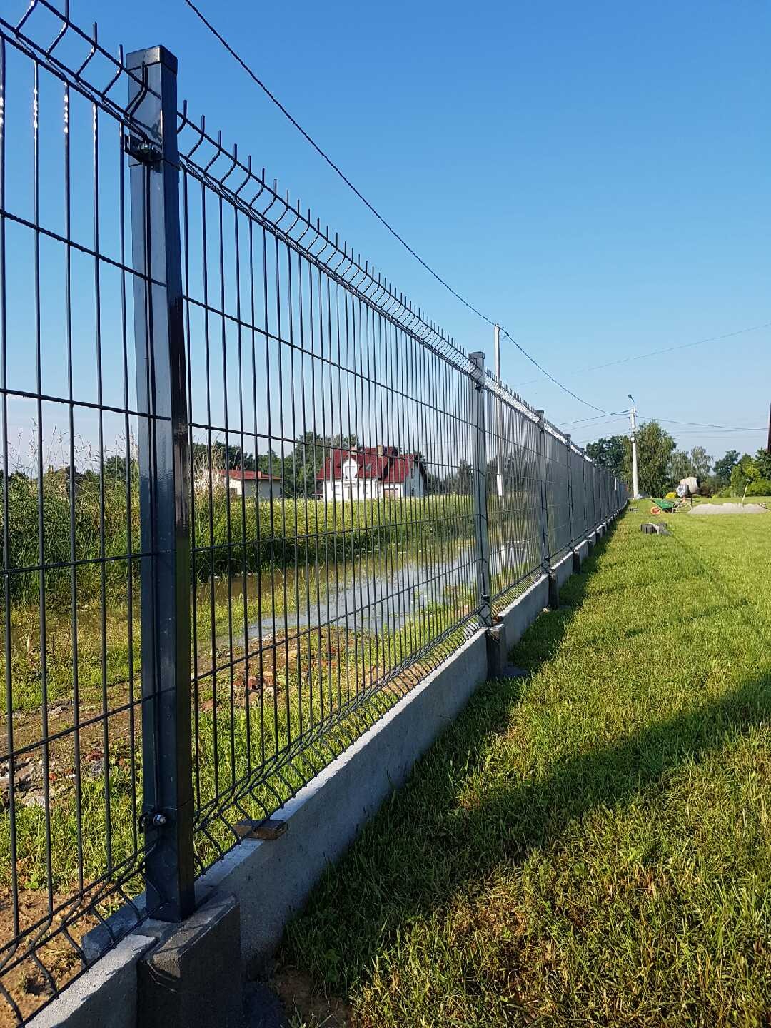 Długi, ciemny panel ogrodzeniowy z siatki, zainstalowany na betonowej podmurówce, rozciągający się wzdłuż granicy posesji z widokiem na rów z wodą i dom w tle pod błękitnym niebem.