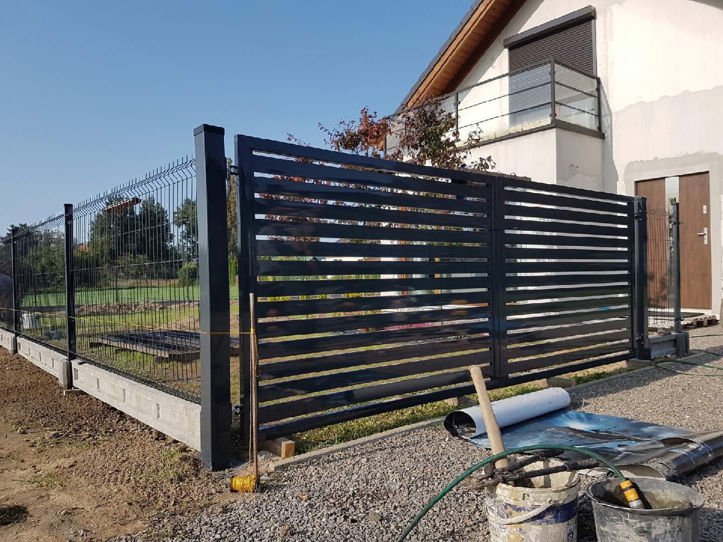 Czarna brama wjazdowa z poziomymi panelami i ogrodzenie panelowe na betonowej podmurówce, widoczne narzędzia i materiały montażowe na żwirowym podłożu, dom w tle.