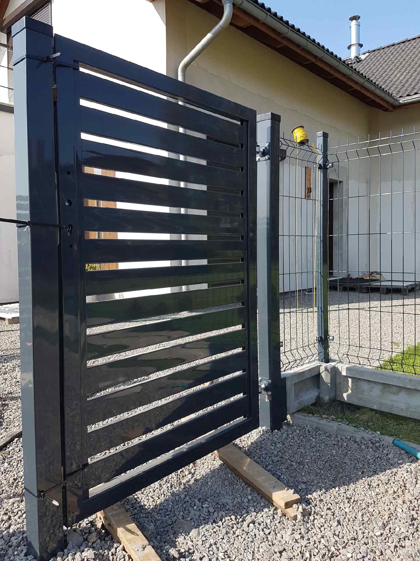 Nowoczesna, pozioma brama wjazdowa z metalowymi lamelami w kolorze antracytowym, zamontowana na słupkach, widoczna na tle ogrodzenia panelowego i domu.