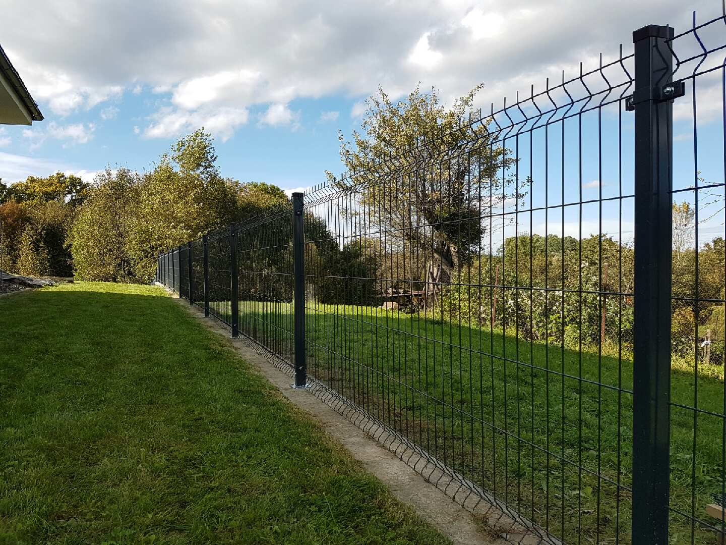Ciemnoszare ogrodzenie panelowe z podmurówką betonową na nierównym terenie, widok z zewnątrz posesji, w tle drzewa i błękitne niebo z chmurami.