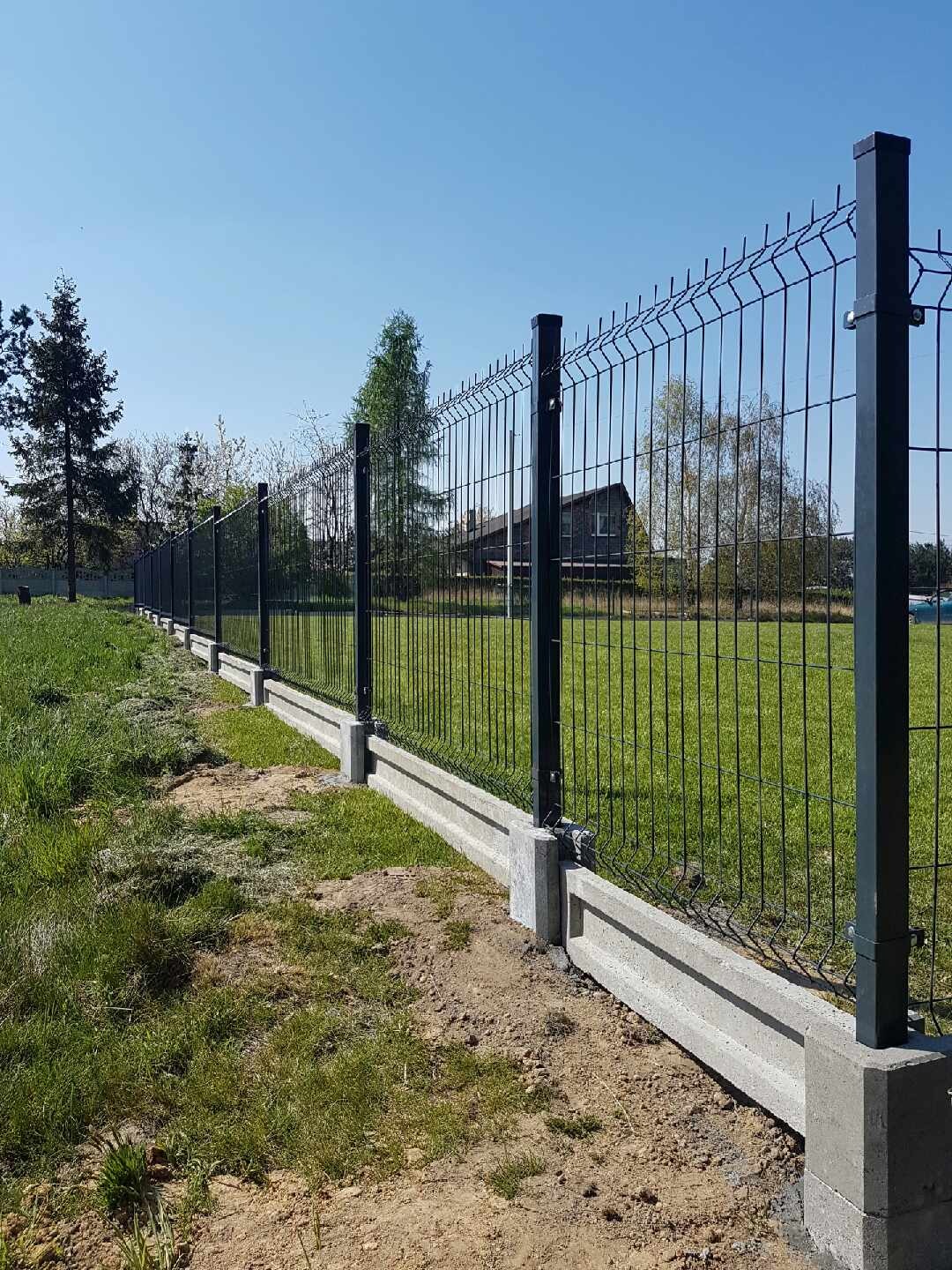 Długie, czarne ogrodzenie panelowe z betonową podmurówką na trawiastym terenie w słoneczny dzień.