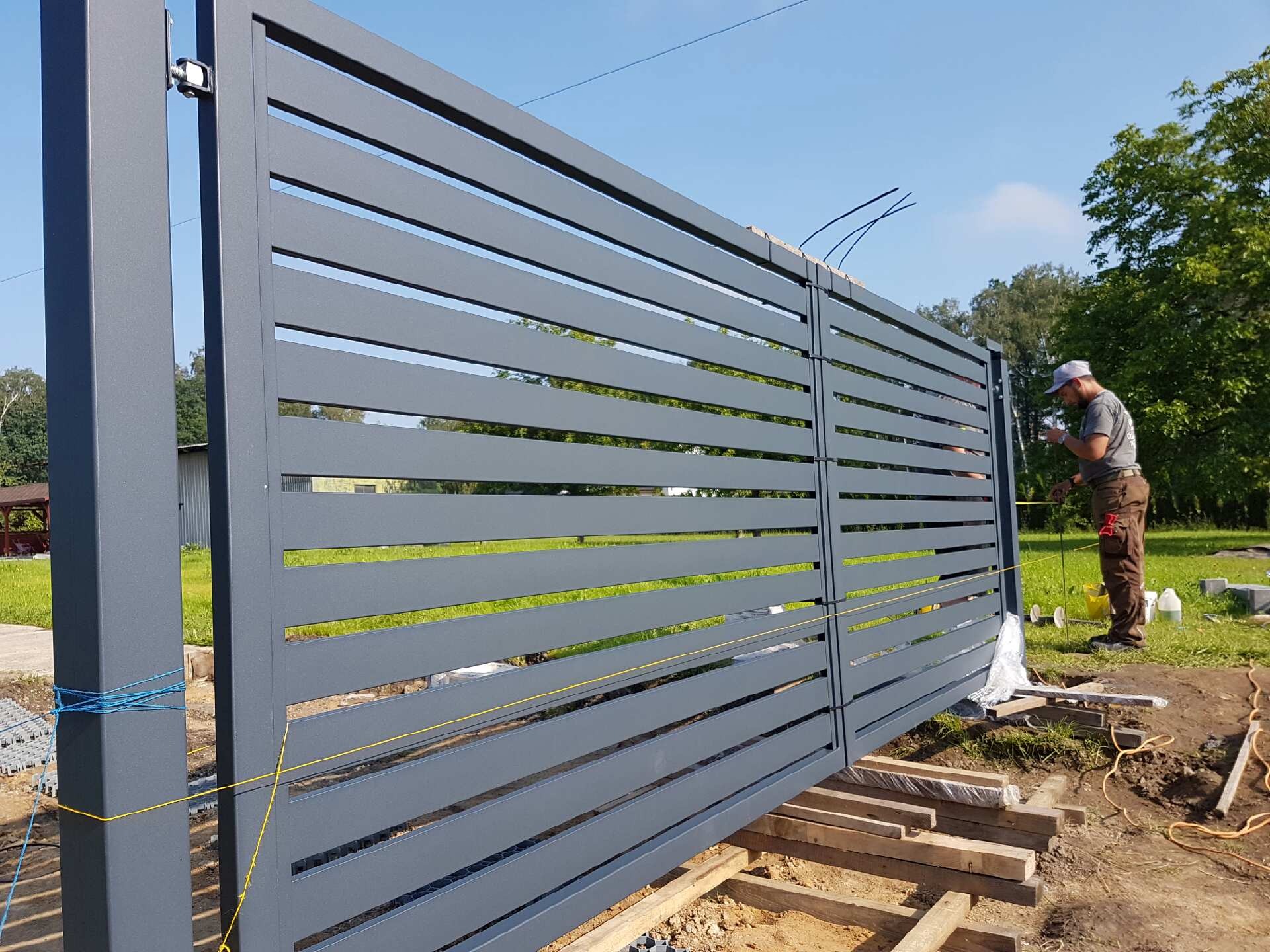 Nowoczesna, przesuwna brama z poziomych, szarych profili metalowych w trakcie montażu; widoczne drewniane podpory, linki pomiarowe i pracownik z miarką.
