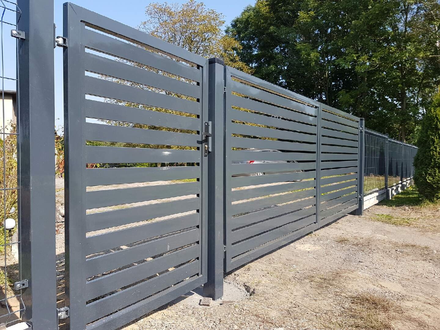 Nowoczesna, przesuwna brama wjazdowa z poziomych, szarych profili, widoczny fragment ogrodzenia panelowego w tle, na podłożu żwirowym.
