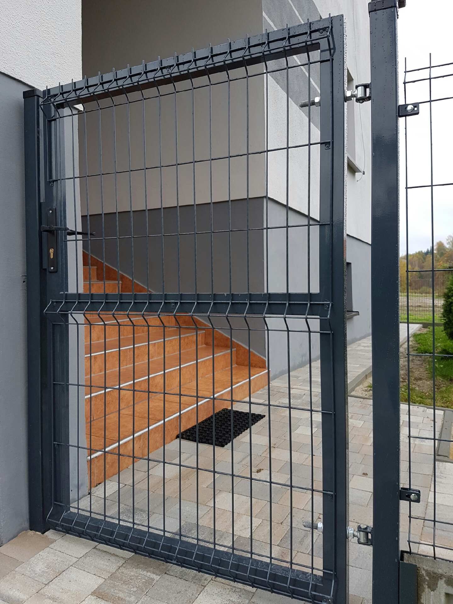 Szara furtka panelowa z zamkiem i widocznymi schodami wejściowymi do budynku