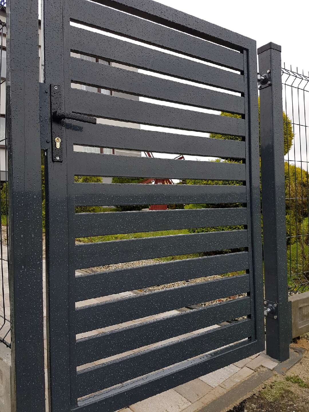 Nowoczesna, antracytowa brama wjazdowa z poziomymi szczeblami, pokryta kroplami deszczu, z widocznym zamkiem i klamką, zamontowana na tle ogrodzenia panelowego i zieleni.