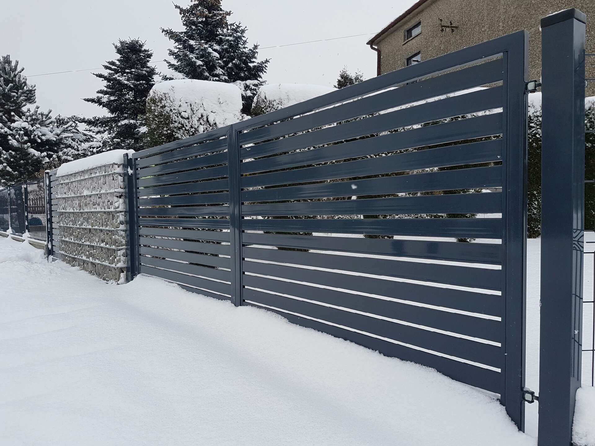 Nowoczesna, pozioma brama wjazdowa z metalowymi panelami w kolorze grafitowym, częściowo zasypana śniegiem, widoczna z zewnątrz posesji w zimowy dzień.
