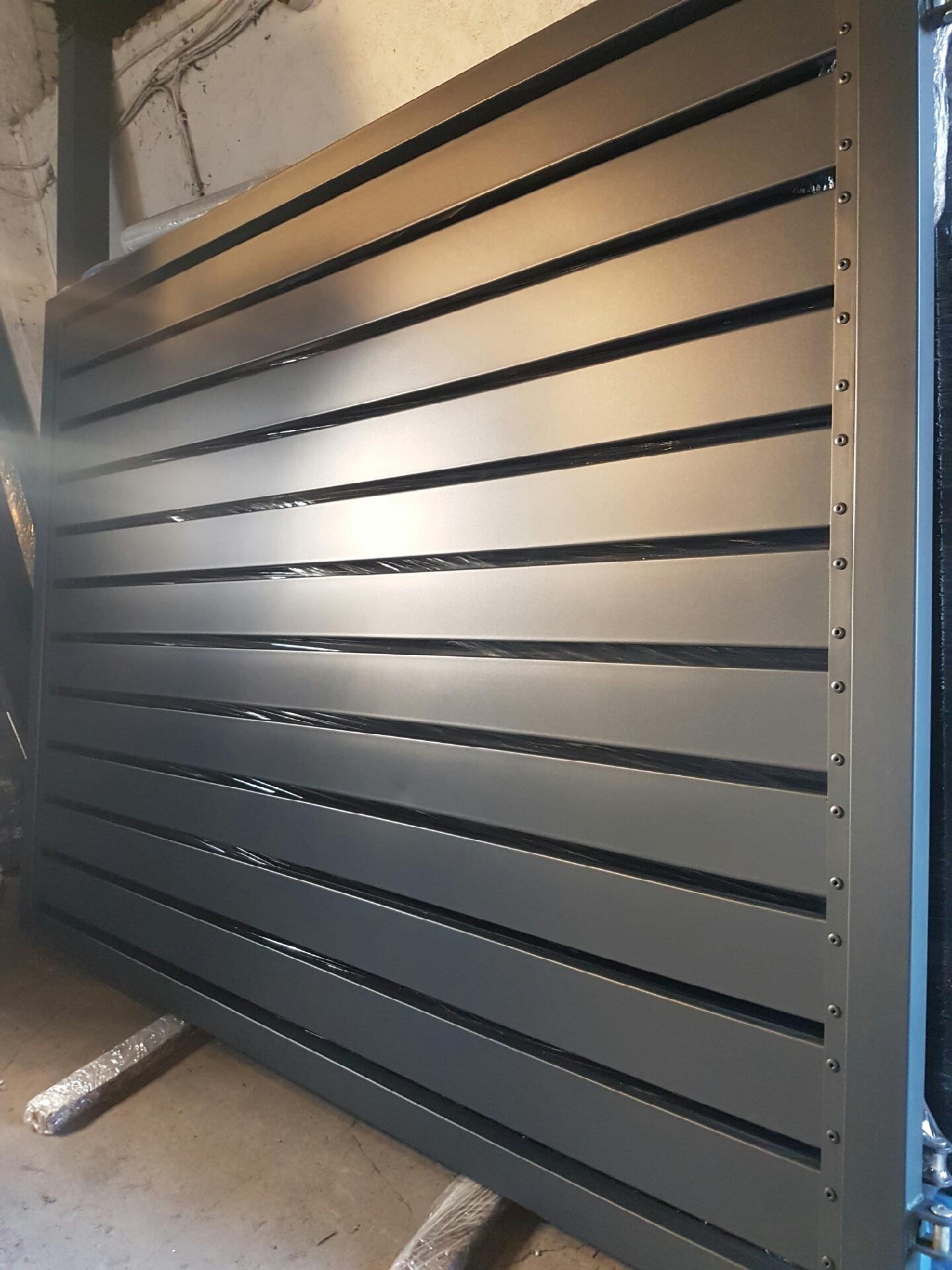 Szara, pozioma brama lamelowa z widocznymi śrubami mocującymi, zabezpieczona folią na podłodze w trakcie transportu lub montażu.