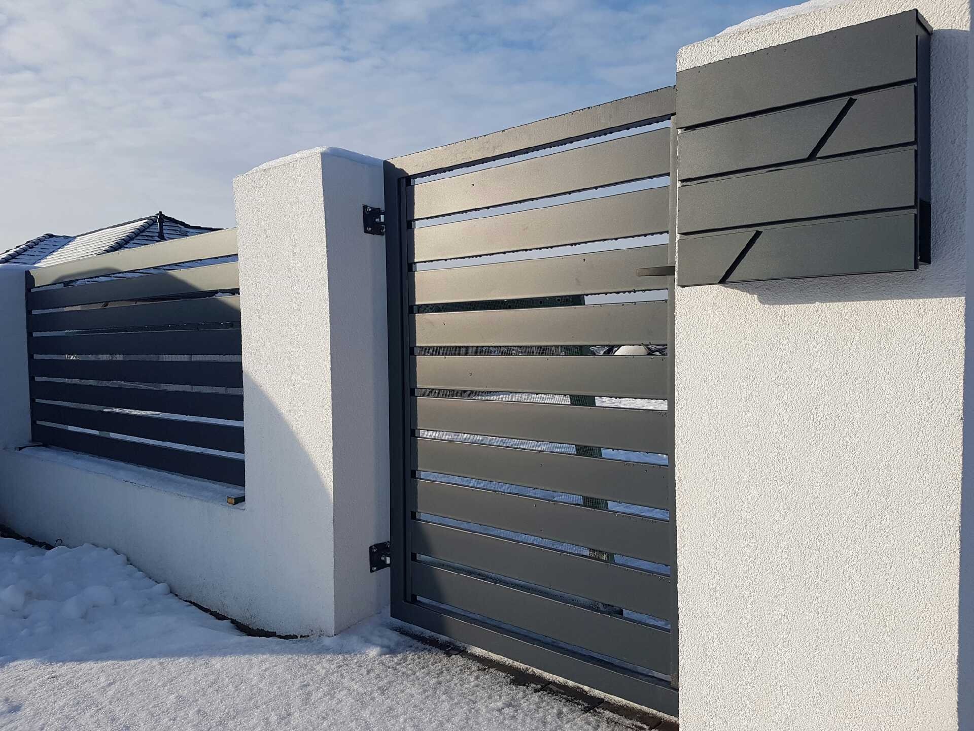 Nowoczesna, pozioma brama wjazdowa z metalowymi panelami w kolorze grafitowym, zamontowana na białych, otynkowanych słupkach. Po prawej stronie, na słupku, skrzynka na listy z geometrycznym wzorem.