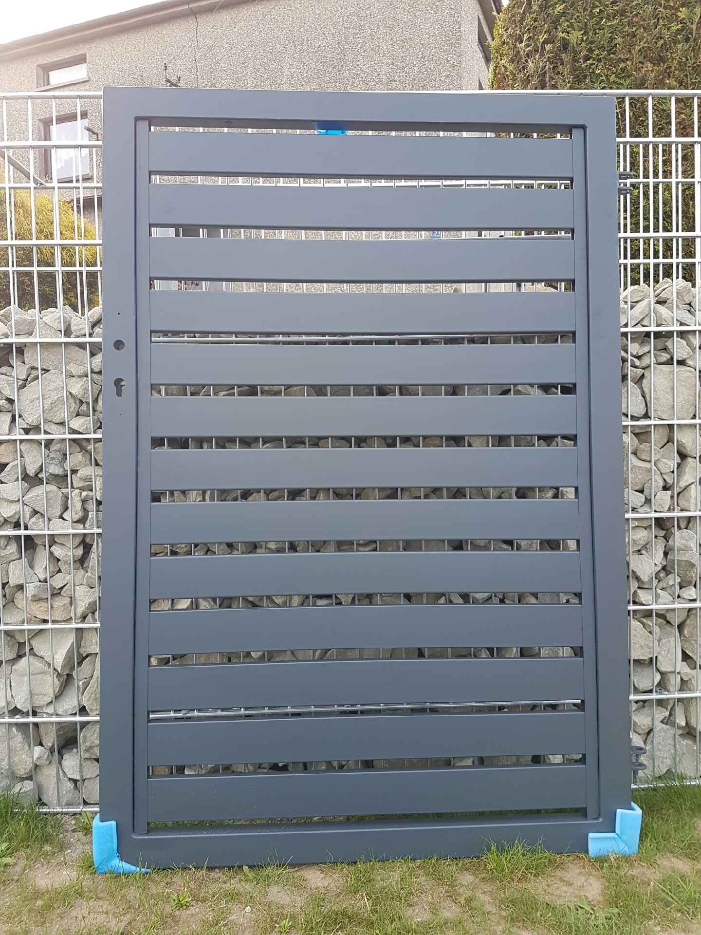 Szara, pozioma brama lamelowa z widocznym zamkiem, oparta na niebieskich narożnikach ochronnych, na tle ogrodzenia gabionowego z kamieniami.