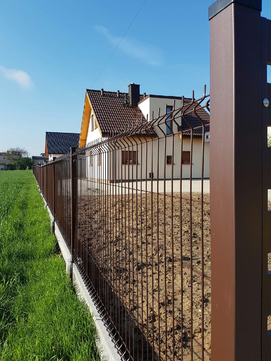 Ciemnobrązowe ogrodzenie panelowe z podmurówką betonową na tle domu jednorodzinnego i błękitnego nieba.