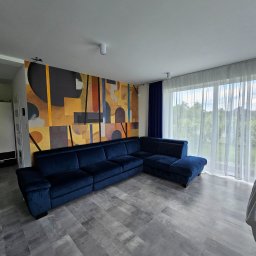 TOM LUX HOME TOMASZ HETNAR - Salon z narożnikiem w odcieniach granatu, parkiet w szarościach i abstrakcyjna tapeta w ciepłych barwach. Duże okno z firanami wpuszcza naturalne światło.