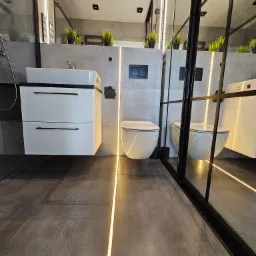Nowoczesna łazienka z szarą podłogą i ścianami, podświetleniem LED, wiszącą białą szafką z umywalką i toaletą typu Geberit, oddzielona czarną, przeszkloną ścianką działową.