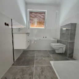 Nowoczesna łazienka w jasnych kolorach z szarymi płytkami podłogowymi, wisząca toaleta, wanna pod oknem z roletami i umywalka z szafką, widoczne przyłącza wodne.