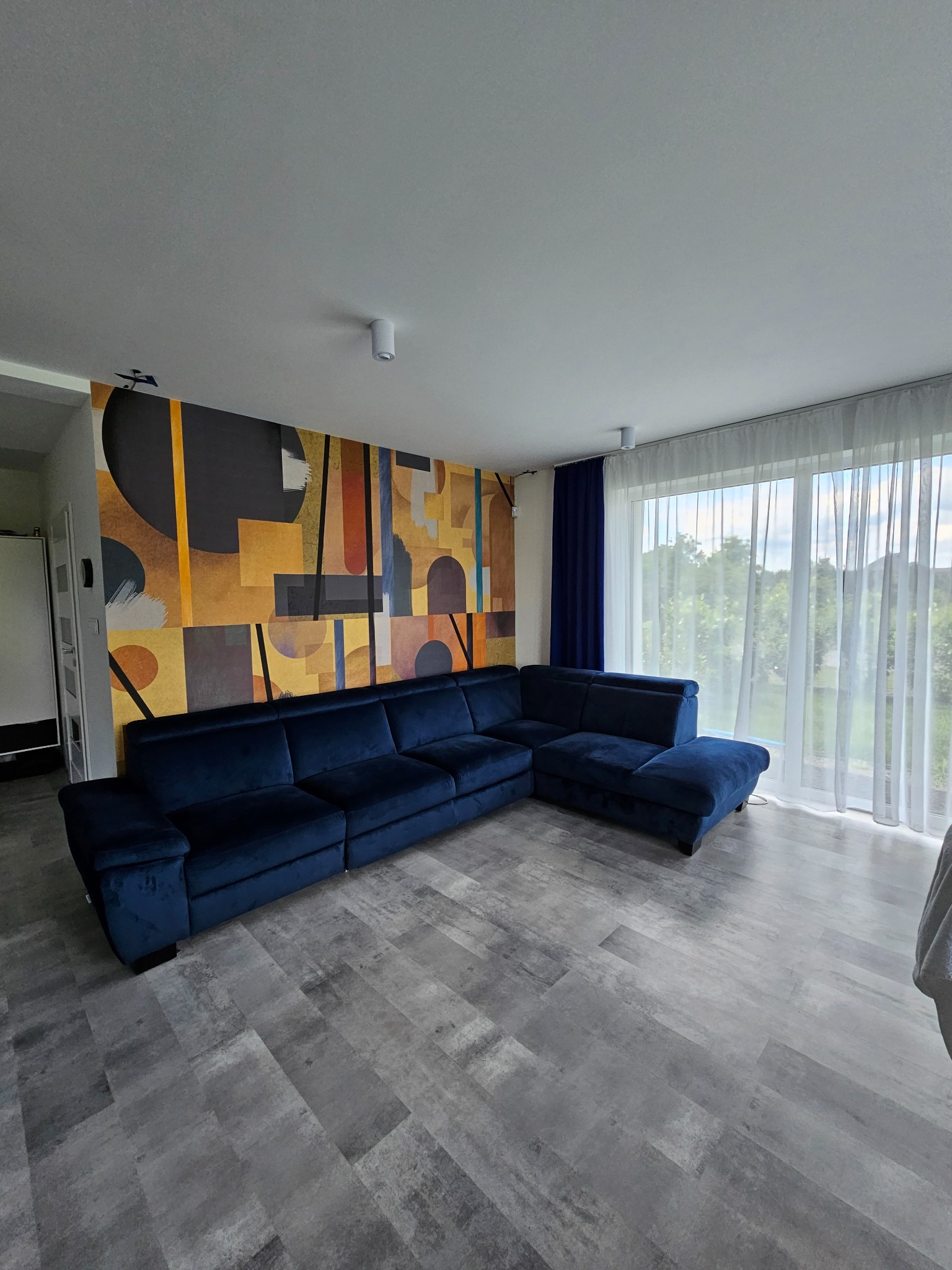 Salon z narożnikiem w odcieniach granatu, parkiet w szarościach i abstrakcyjna tapeta w ciepłych barwach. Duże okno z firanami wpuszcza naturalne światło.