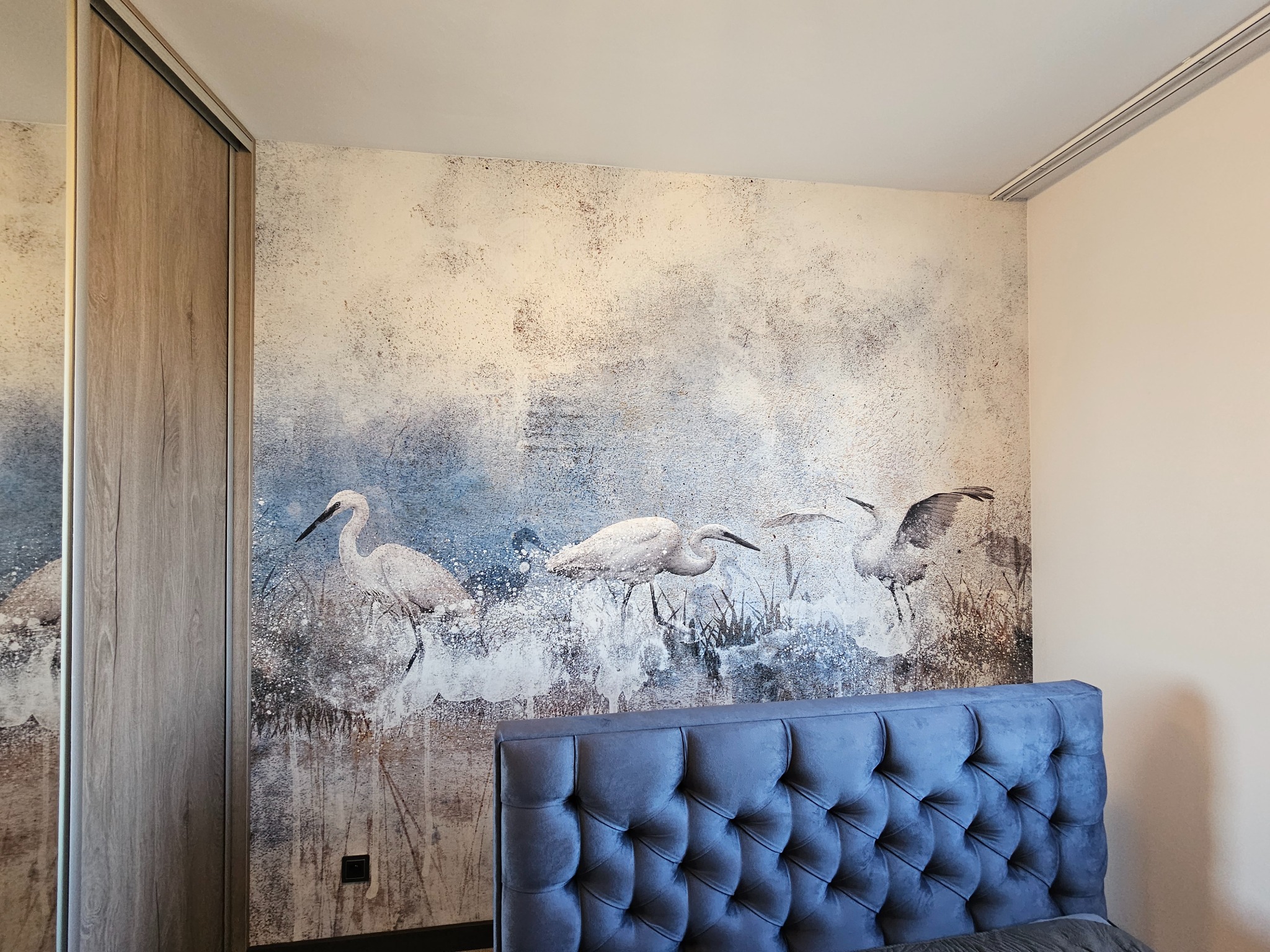 Ściana w sypialni z dekoracyjną tapetą przedstawiającą żurawie, widoczny fragment łóżka z wezgłowiem i szafa przesuwna. Nowoczesny design wnętrza.