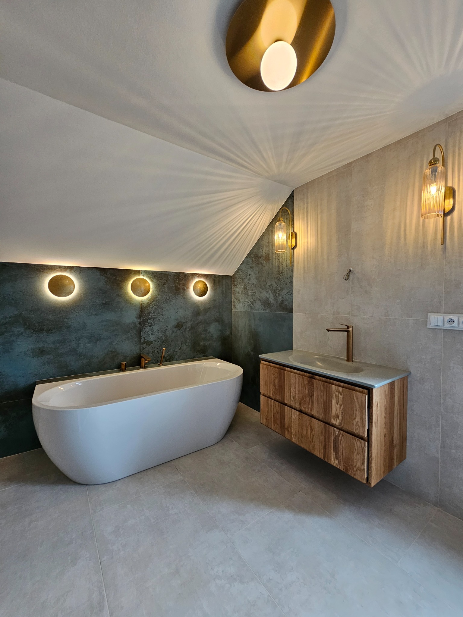 Nowoczesna łazienka na poddaszu z wolnostojącą wanną, drewnianą szafką pod umywalką i oświetleniem punktowym. Szare płytki i akcenty złota.