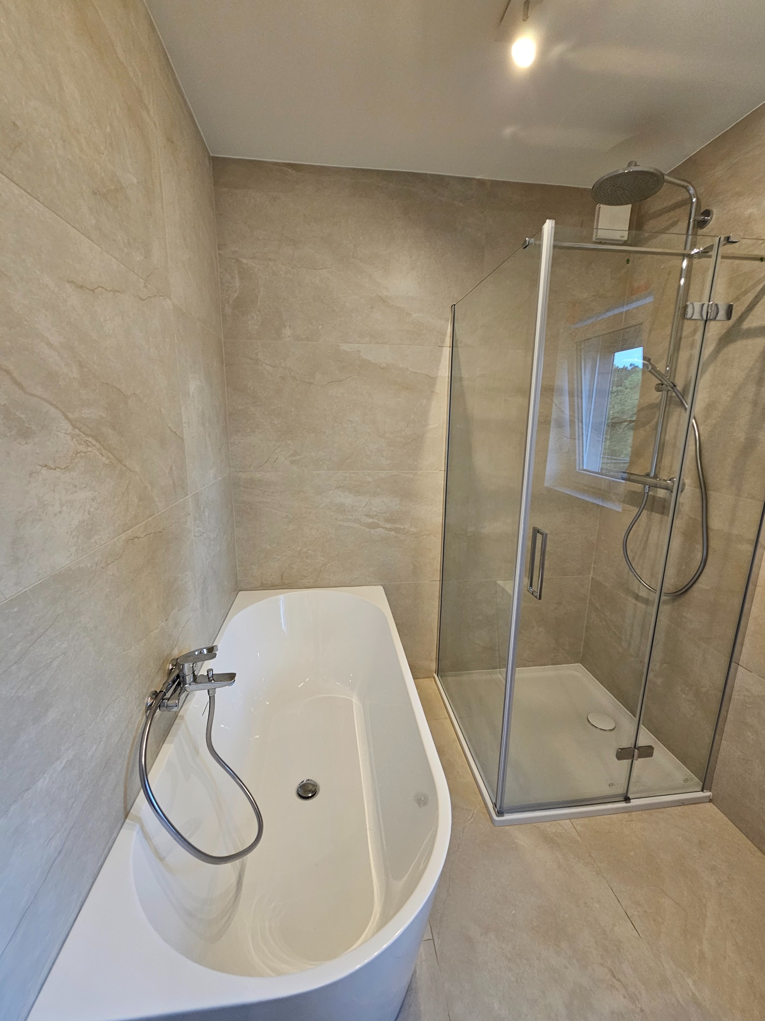 Nowoczesna łazienka z beżowymi płytkami imitującymi kamień, białą wanną i kabiną prysznicową ze szklanymi drzwiami. Minimalistyczny design, jasne oświetlenie.