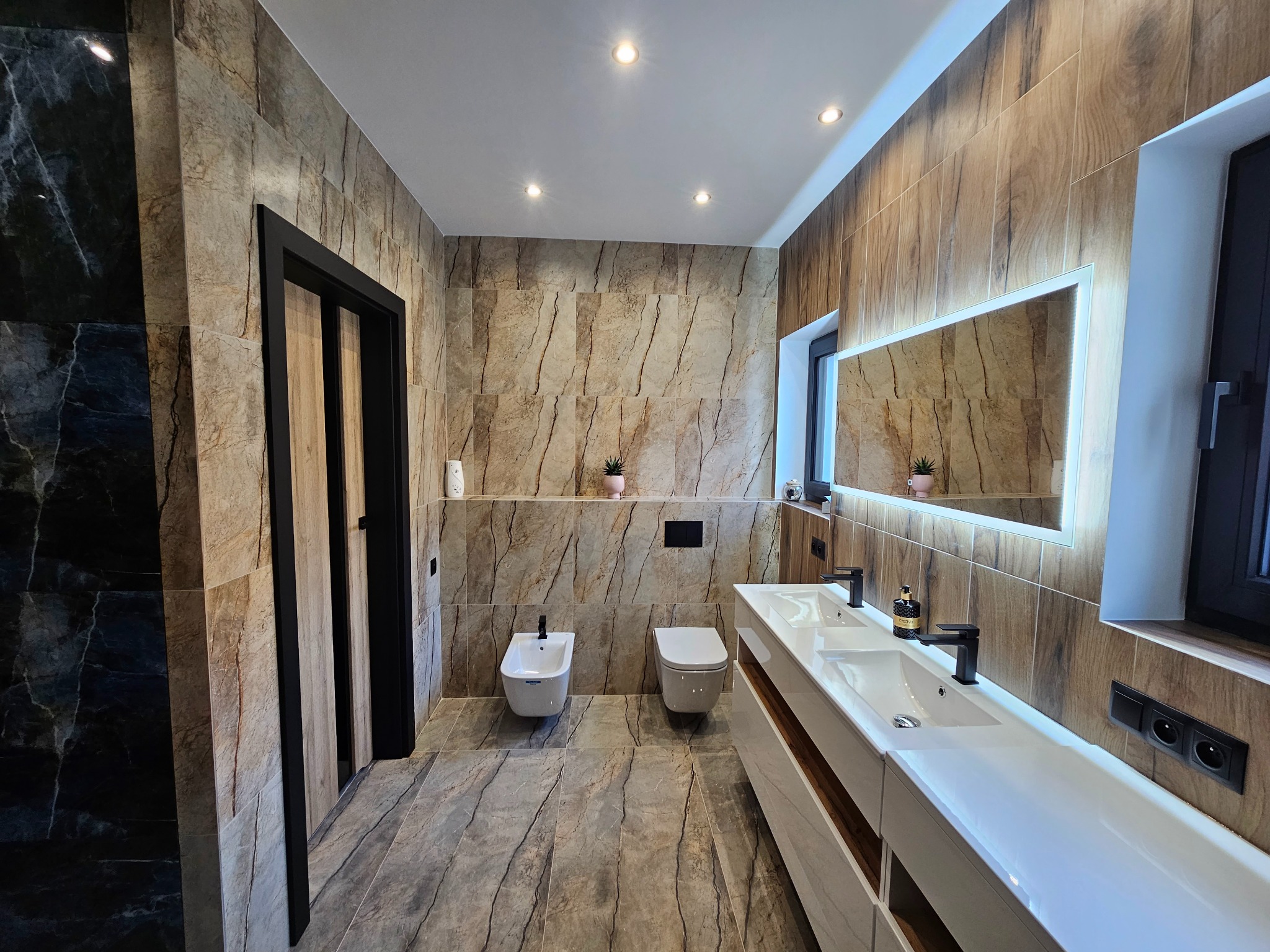 Nowoczesna łazienka z marmurowymi płytkami imitującymi drewno, podświetlanym lustrem i czarnymi akcentami. Bidet i toaleta w minimalistycznym stylu.