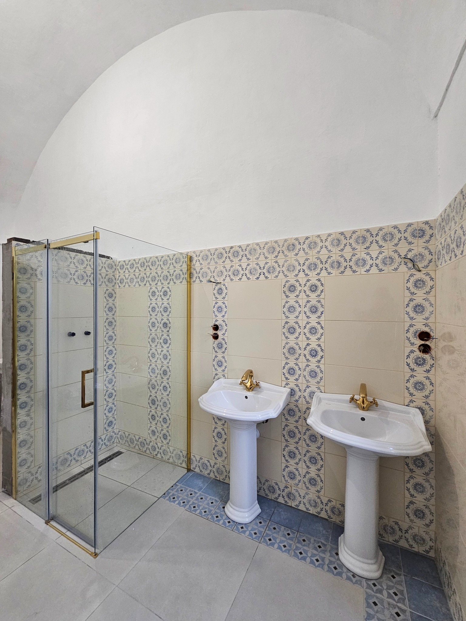 Wykończona łazienka z kabiną prysznicową w złotej ramie i dwiema umywalkami na postumentach. Płytki w kremowo-niebieskie wzory. Sufit sklepiony.