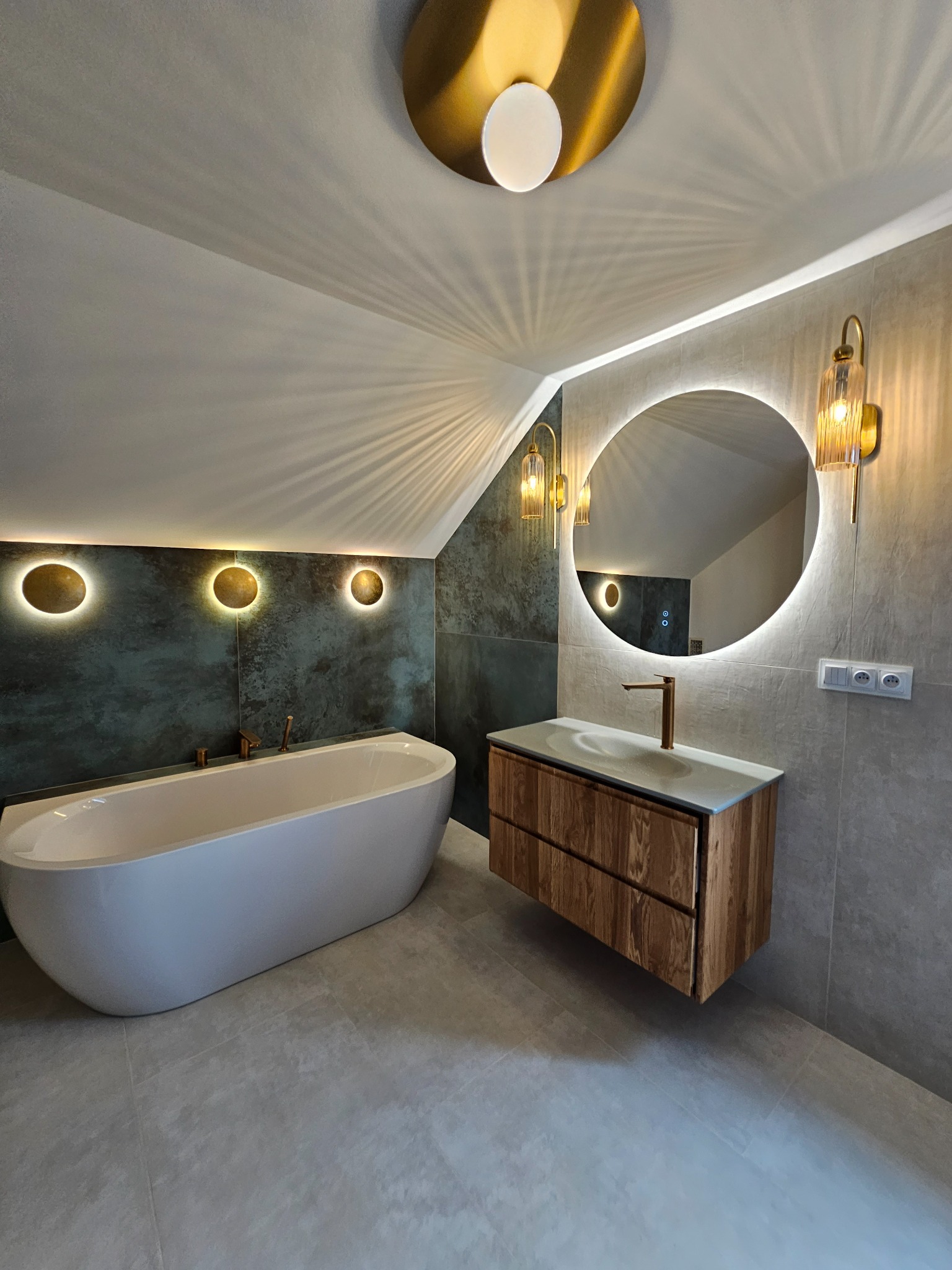 Nowoczesna łazienka na poddaszu z wanną, umywalką z drewnianą szafką i okrągłym lustrem z podświetleniem LED. Złote akcenty oświetleniowe.