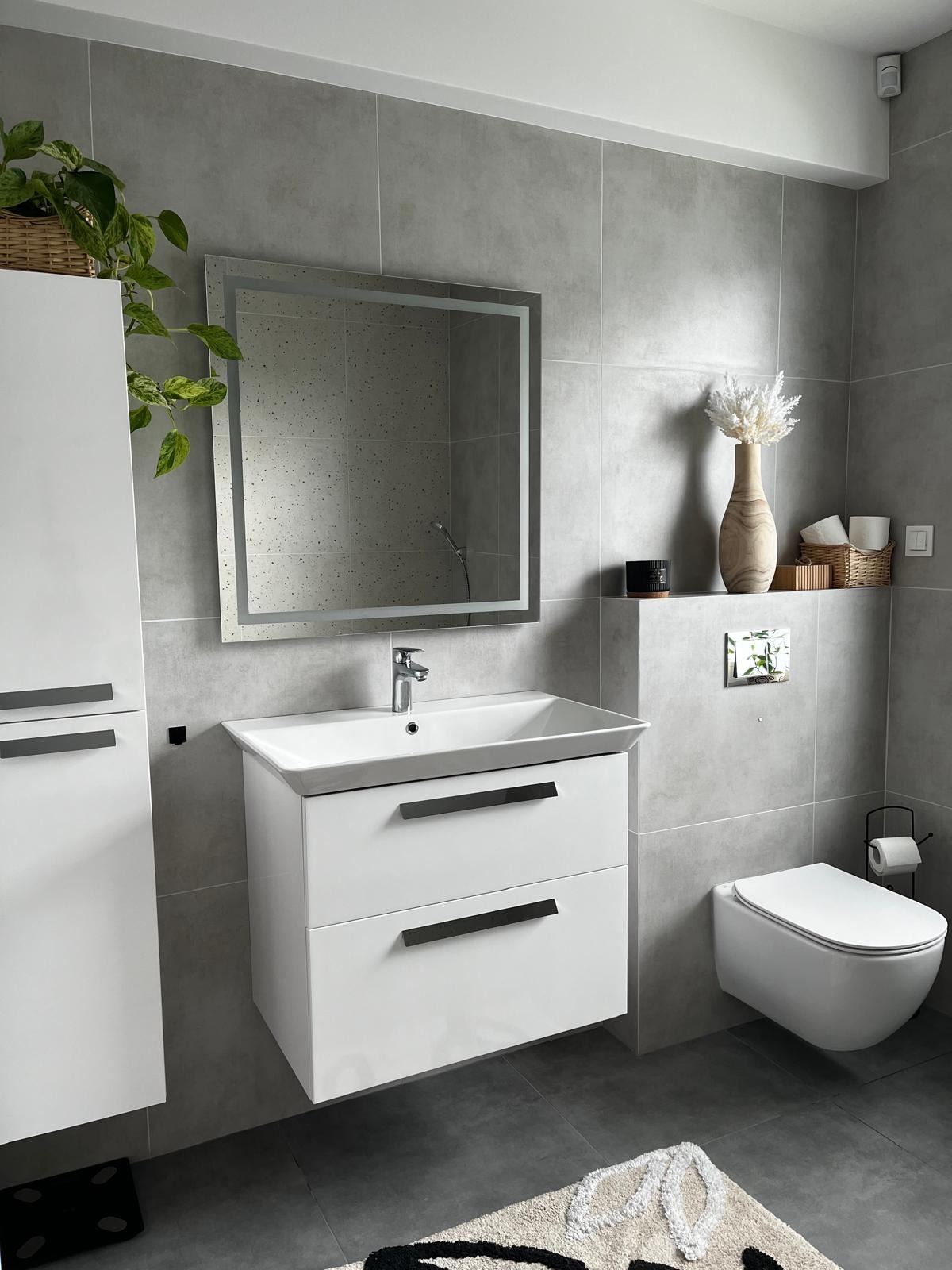 Nowoczesna łazienka w szarościach z białymi meblami: szafka pod umywalkę z dwiema szufladami, podświetlane lustro i wisząca toaleta. Uzupełniona o rośliny i dekoracje.