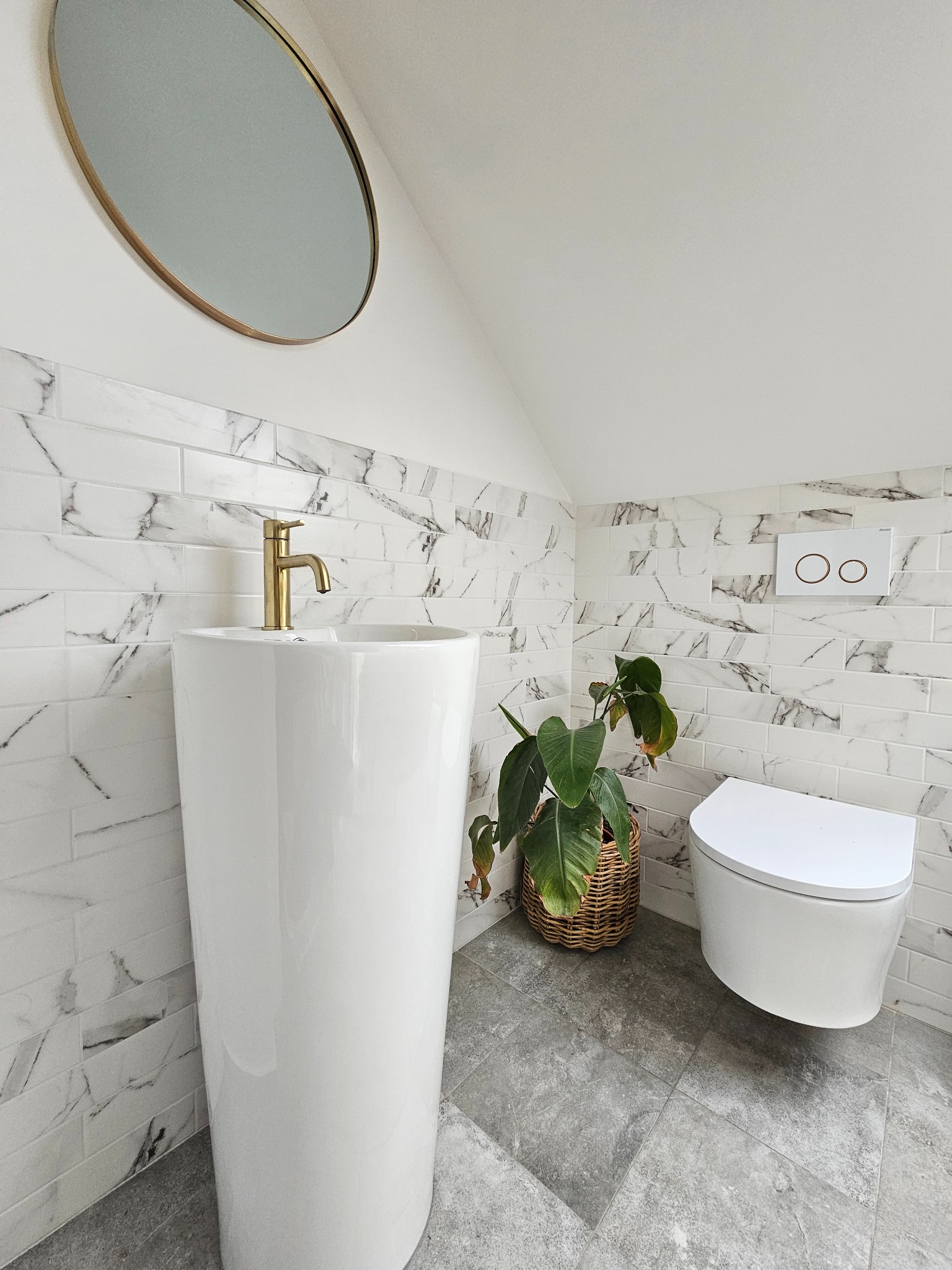 Nowoczesna łazienka na poddaszu: biała umywalka kolumnowa z złotą armaturą, marmurowe płytki, wisząca toaleta i okrągłe lustro w złotej ramie.