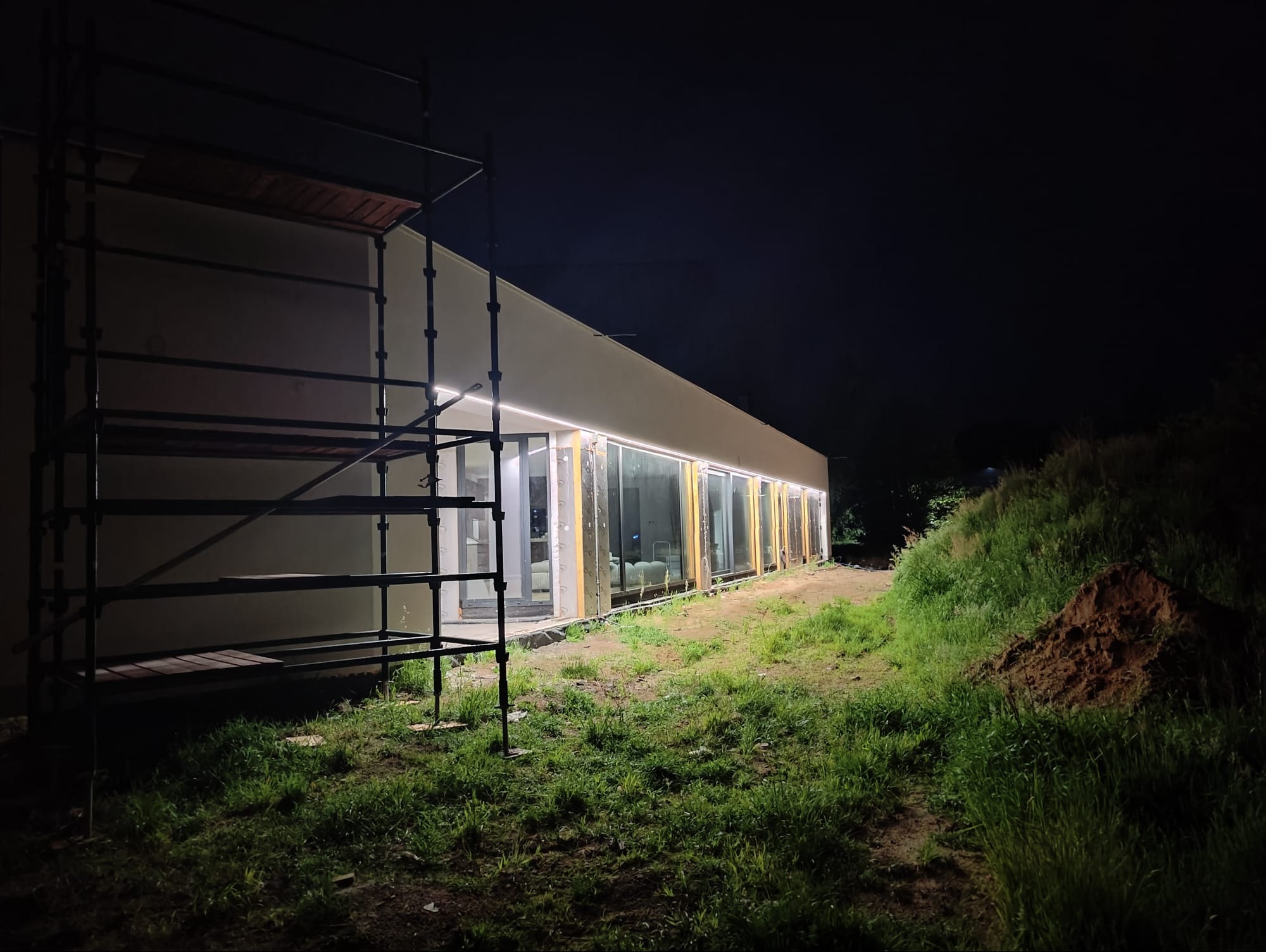 Nocny widok budynku w trakcie budowy z zamontowanym oświetleniem zewnętrznym w postaci taśmy LED, widoczne rusztowanie i niezagospodarowany teren.