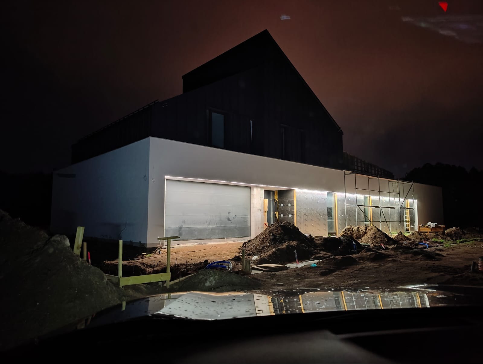Nowoczesny dom jednorodzinny w trakcie wykończenia, nocą, z zamontowanym zewnętrznym oświetleniem taśmą LED, widoczne rusztowania i przygotowany teren.