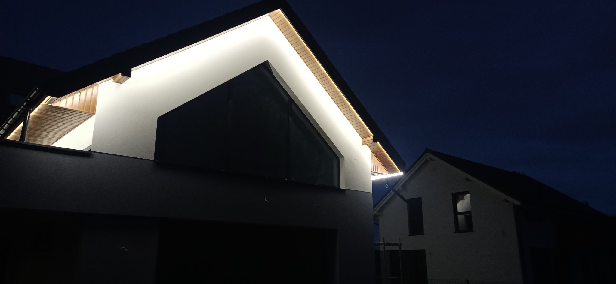 Nowoczesny dom jednorodzinny oświetlony zewnętrzną taśmą LED, widoczne detale drewnianego podbitki dachowej, nocne ujęcie.
