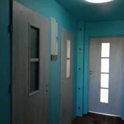 Korytarz w turkusowym kolorze z trzema drewnopodobnymi drzwiami z przeszkleniami, widoczna skrzynka elektryczna na ścianie i brązowa podłoga z płytek.