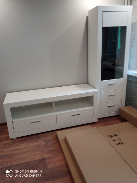 Biały zestaw mebli pokojowych: szafka RTV z szufladami i półkami oraz wysoka witryna z przeszklonym frontem, częściowo rozpakowane kartony na podłodze.