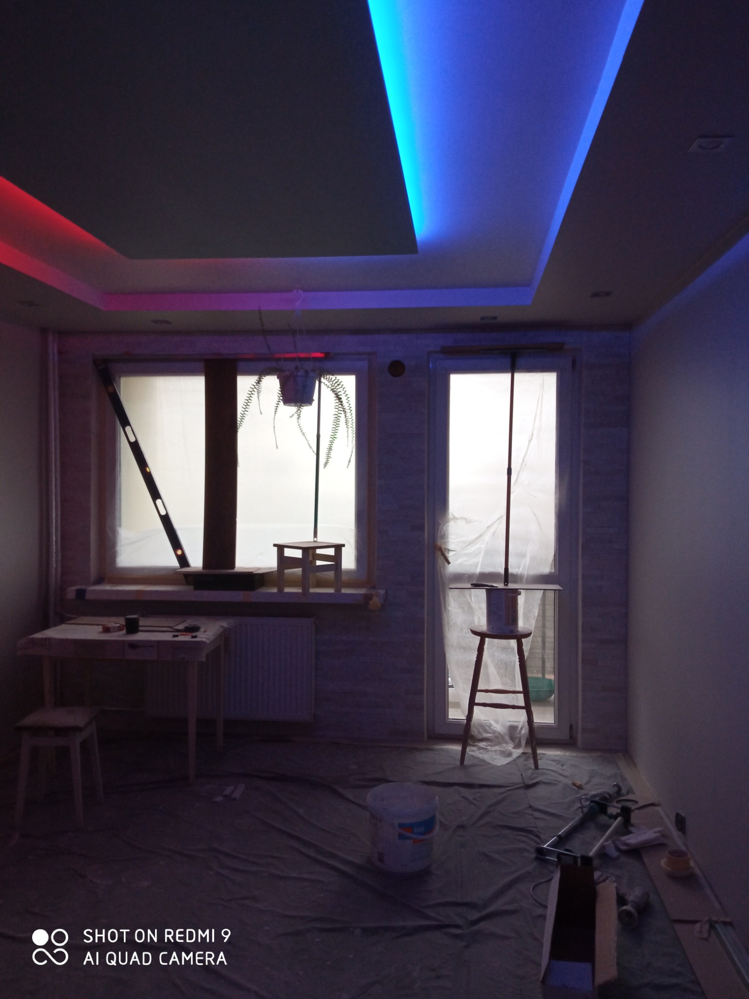 Pokój w trakcie prac wykończeniowych: sufit z oświetleniem LED w kolorach niebieskim i czerwonym, okna zabezpieczone folią, podłoga przykryta folią ochronną, widoczne narzędzia malarskie i drabina.