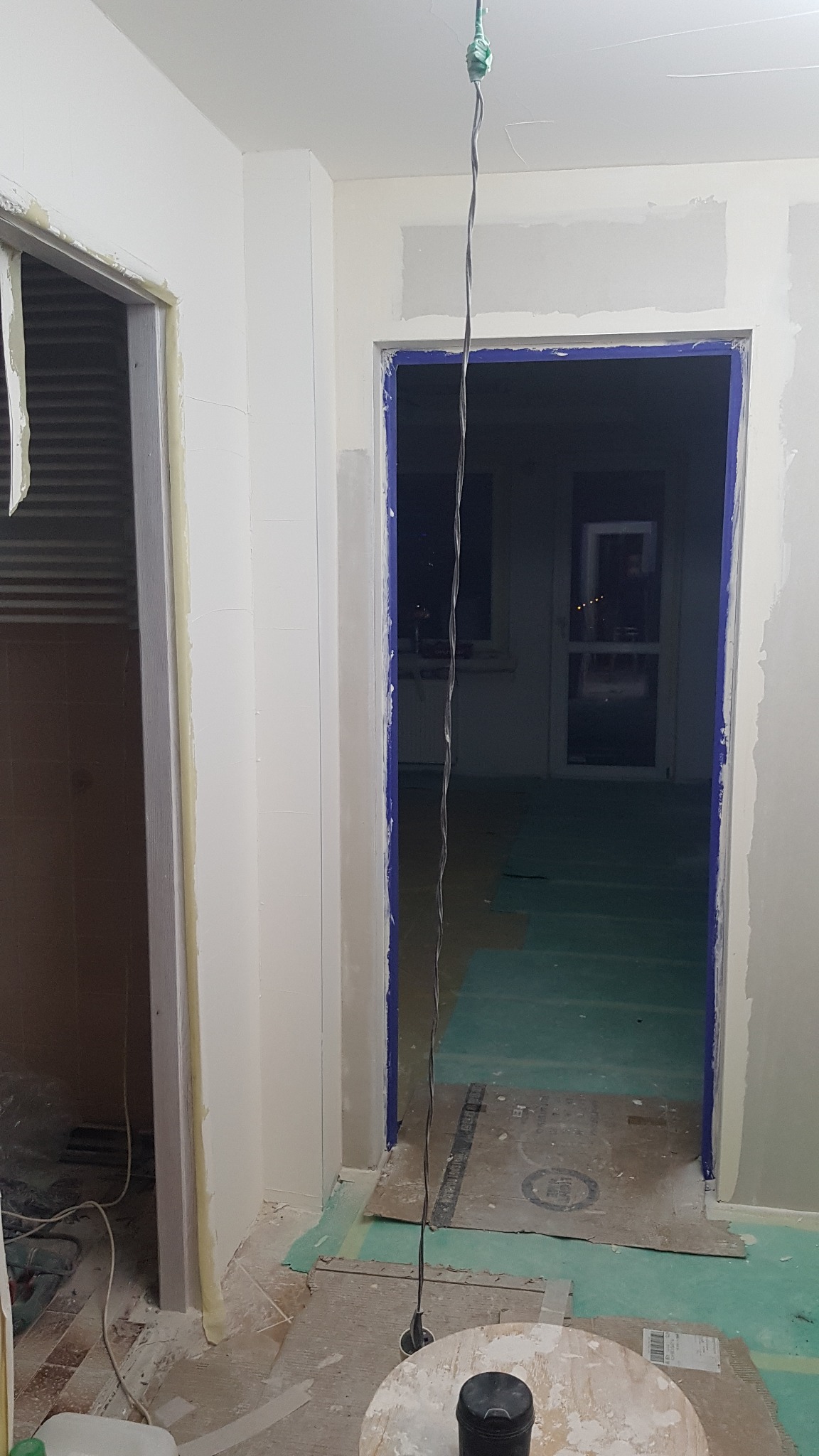 Przygotowanie do malowania ścian w remontowanym mieszkaniu: widoczne dwa otwory drzwiowe, jeden zabezpieczony taśmą malarską, drugi z niebieską folią ochronną, podłoga zabezpieczona kartonem...