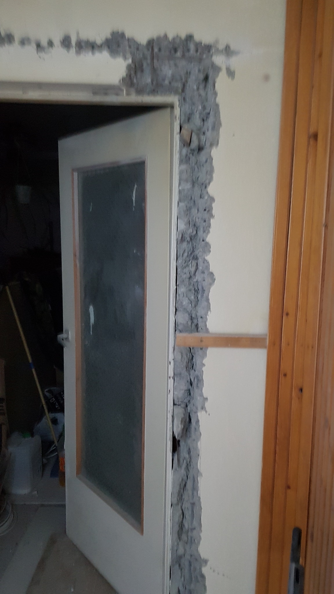 Przygotowanie ościeżnicy do montażu drzwi: surowa ściana z nierównomiernie nałożoną zaprawą wyrównującą, widoczny fragment drewnianej listwy i częściowo otwarte drzwi z szybą.