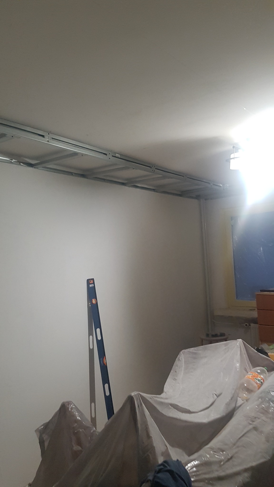 Wnętrze w trakcie prac przygotowawczych: profile sufitowe zamontowane, ściany przygotowane, meble zakryte folią ochronną, poziomica oparta o ścianę.