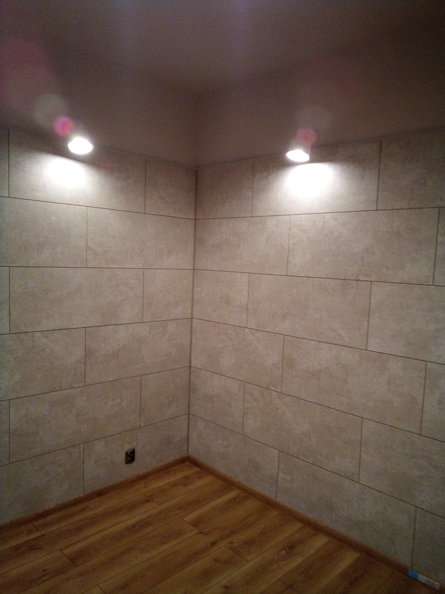 Kąt w pomieszczeniu wyłożonym beżowymi, prostokątnymi płytkami ceramicznymi, z drewnianą podłogą i dwoma punktowymi lampami sufitowymi