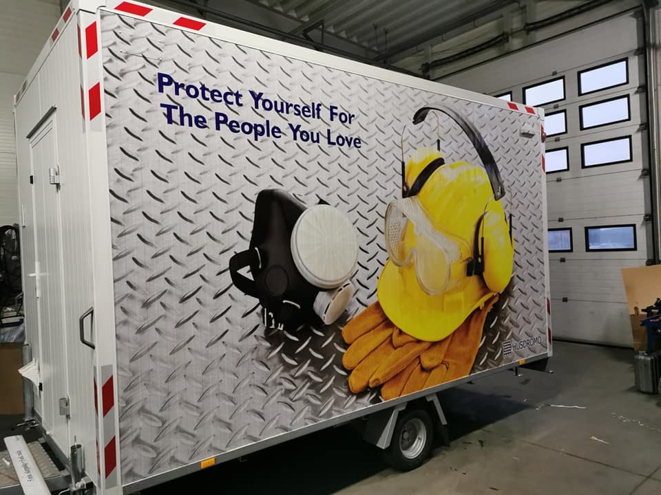 Przyczepa z oklejoną grafiką BHP, prezentująca maskę, kask, okulary ochronne i rękawice na tle metalowej blachy ryflowanej oraz hasło 'Protect Yourself For The People You Love'.