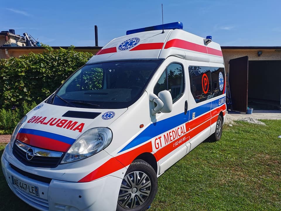 Oklejony ambulans GT Medical z czerwono-niebieskimi pasami na białym tle, zaparkowany na trawie przed budynkiem.